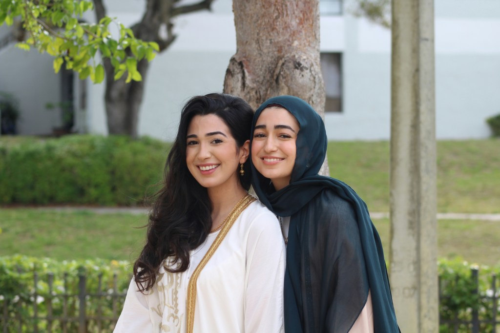 two young women who appear to be sisters, one with hijab, the other with long wavy black hair, pose in front of a tree trunk